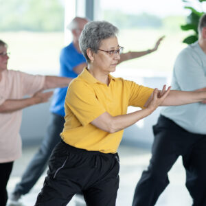 A small group of seniors are seen practicing Tai Chi in an indoor studio. They are each dressed comfortably and are focused on their movements and proper form as they practice the Martial Art.
