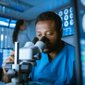 Medical Research Scientist Looking under the Microscope in the Laboratory. Neurologist Solving Puzzles of the Mind and Brain. In the Laboratory with Multiple Screens Showing MRI / CT Brain Scan Images.